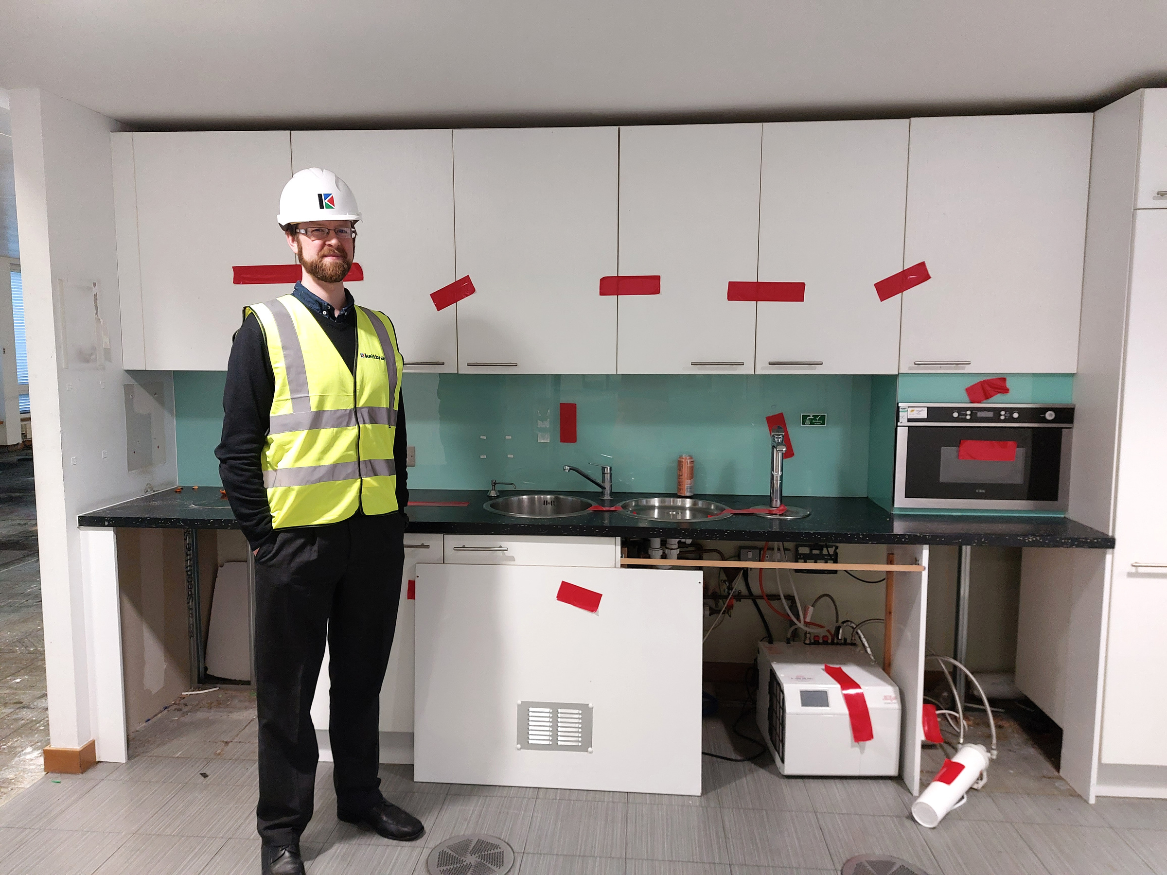 Ed Scolding wearing a hard hat and high-visibility vest beside kitchen fittings marked for removal.