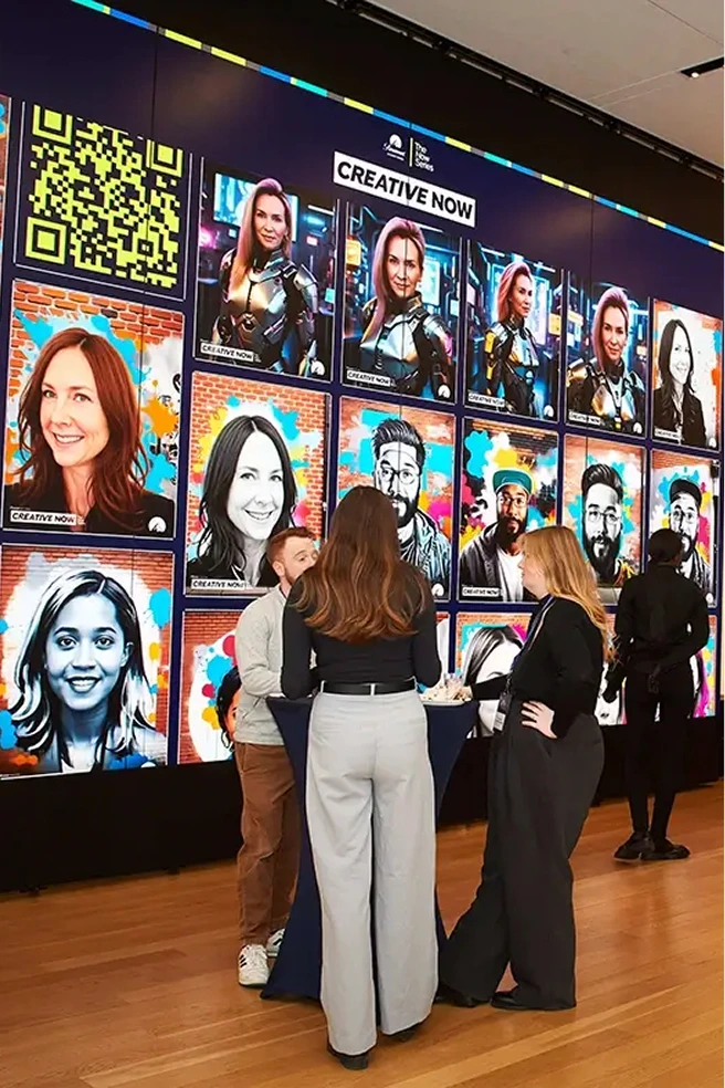 Three people standing and talking in front of a large digital wall displaying colorful portraits labeled 'Creative Now'.