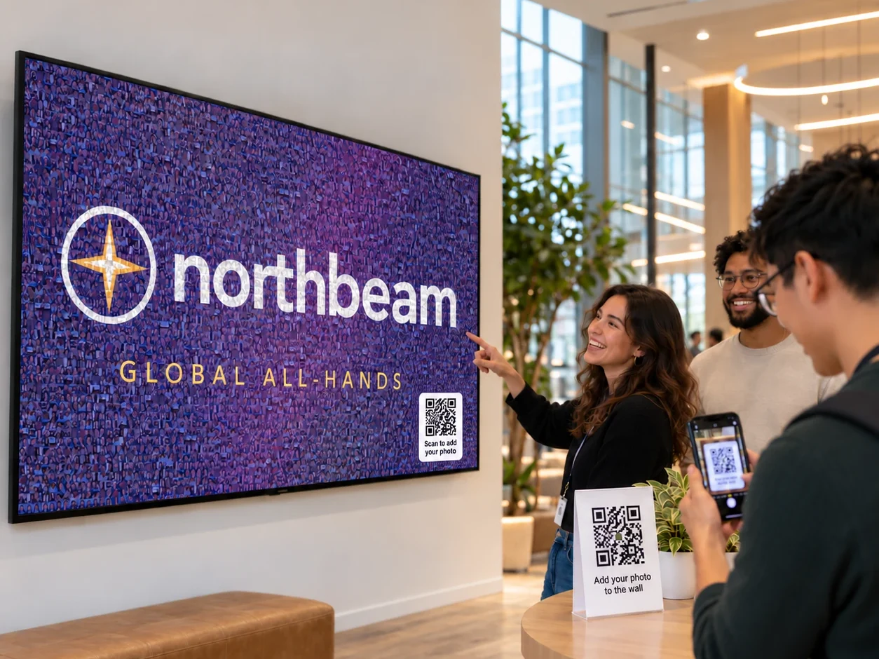Employees in an office lobby scanning a QR code on a table sign to add photos to a live photo mosaic on a wall-mounted screen, with no photo booth