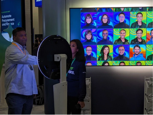 Two people beside a ring light photo kiosk next to a screen gridded with colorful attendee headshots