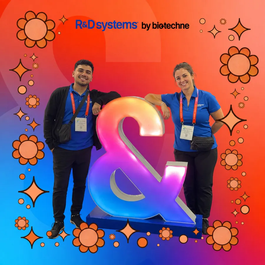 Photo booth snapshot of two teammates leaning on a glowing ampersand statue on an R&D Systems by Bio-Techne frame
