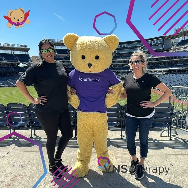 Photo booth snapshot of two guests posing with a teddy bear mascot in a VNS Therapy shirt at a stadium