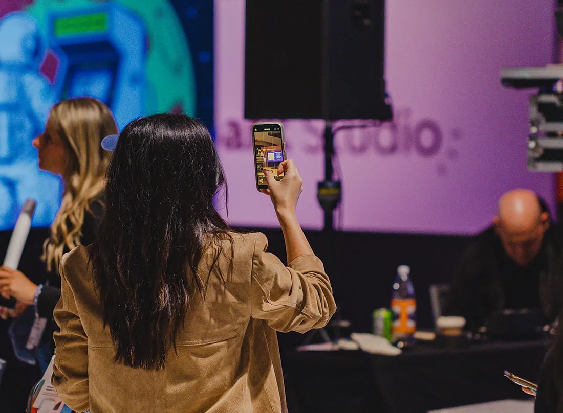 An attendee holds up her phone to record a large event screen while other guests and a staffed table fill the room