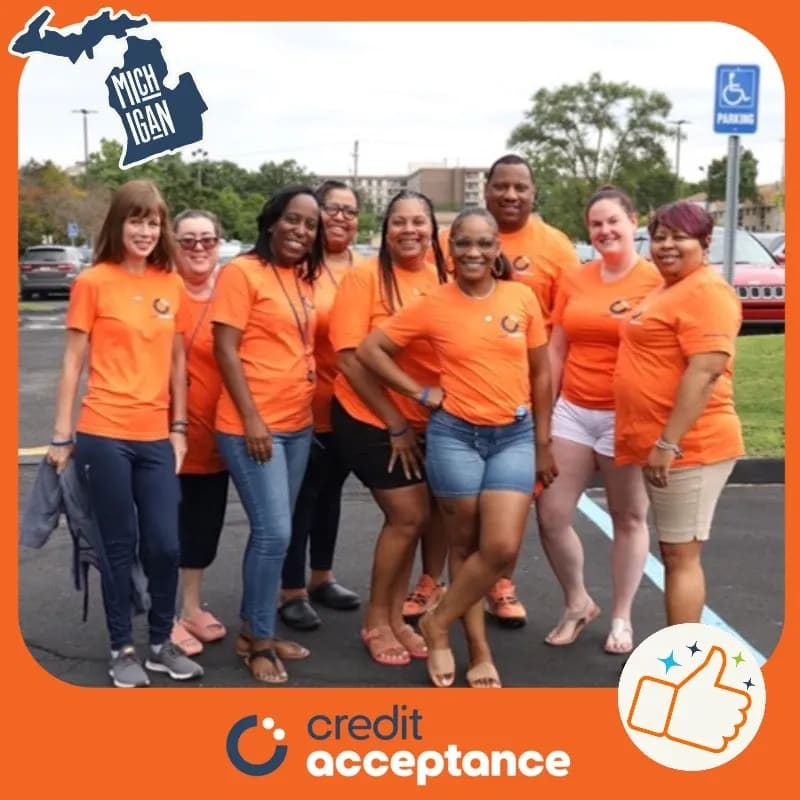 Ten employees in matching orange shirts posing in a parking lot inside an orange Credit Acceptance frame with a Michigan sticker