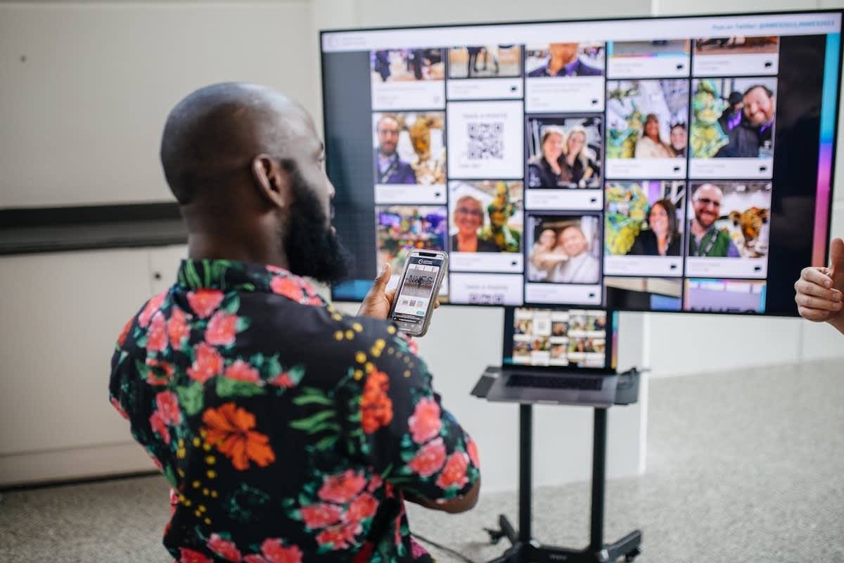 A guest holds up his phone in front of a large screen running a live gallery of photos from the event, with a laptop driving the display below it
