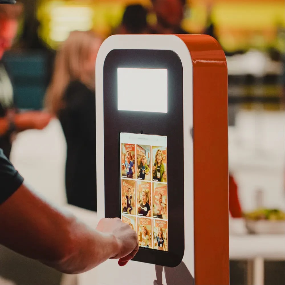 A guest tapping an iPad mounted in an orange and white kiosk stand at a conference, with a grid of illustrated AI portraits on the screen