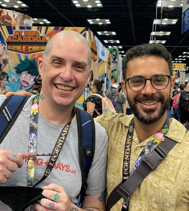 Two smiling men with lanyards at a convention with My Hero Academia posters in the background.