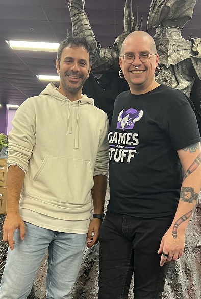 Two men standing and smiling indoors with a large dragon statue behind them.