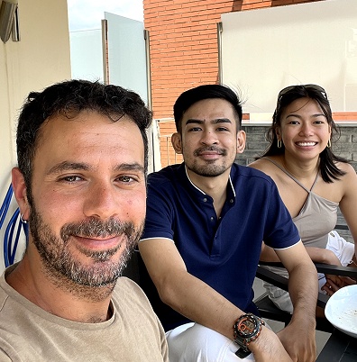 Three people smiling and sitting at an outdoor table with a brick wall in the background.