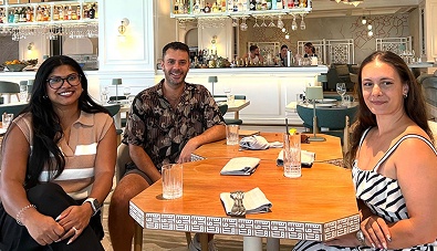 Three people sitting at a wooden table with drinks in a bright restaurant or cafe setting.