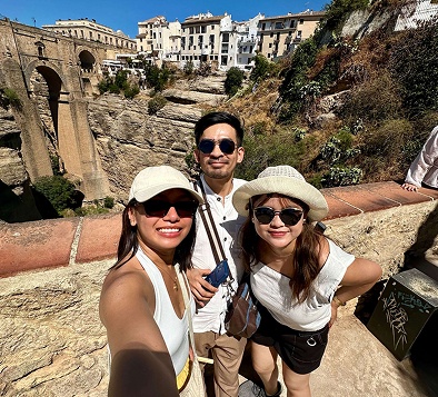 Three friends wearing sunglasses smiling for a selfie near a historic stone bridge and cliffside town.