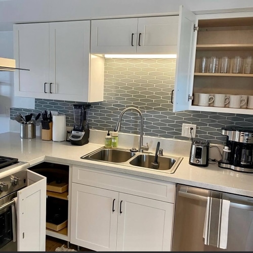 Compact kitchen with white cabinetry, double sink, gray hexagonal tile backsplash, and various small appliances on the countertop.