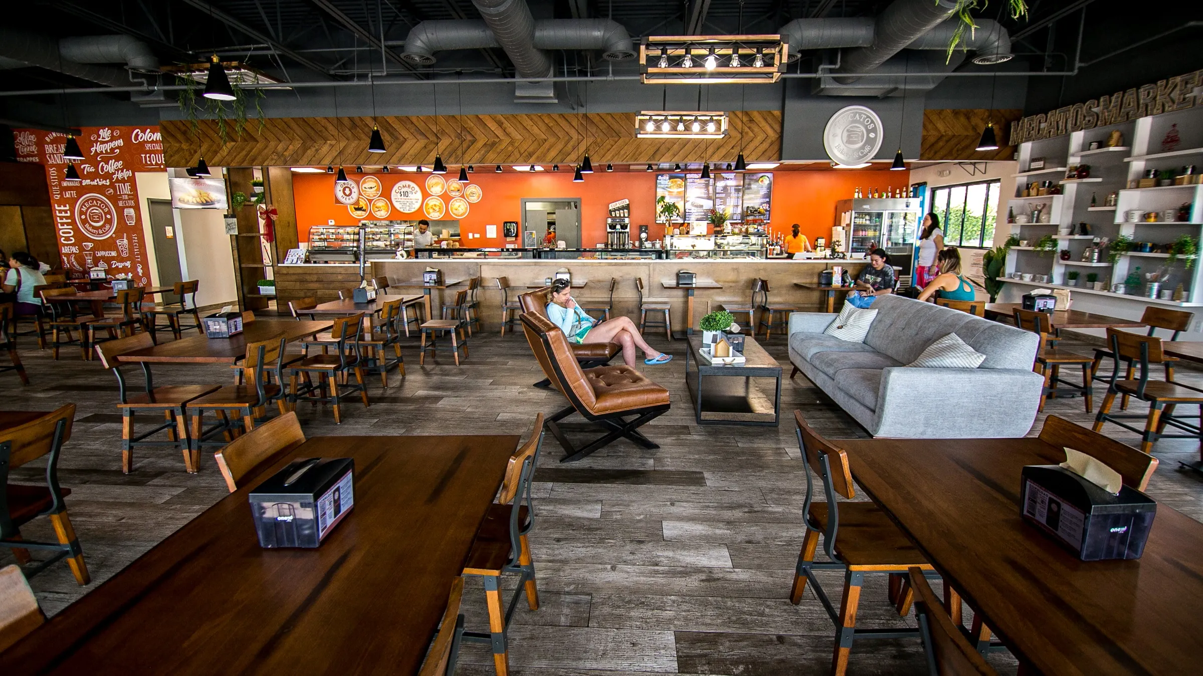 Spacious cafe interior with wooden tables and chairs, brown leather chairs near a gray sofa, and customers at the counter and seating area.