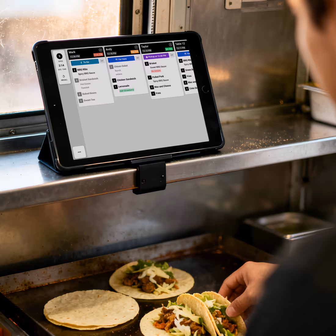 Digital kitchen order screen showing two open orders for Josh and Ashley, with preparation times, meal priority, and item details including modifications and crossed-out items.