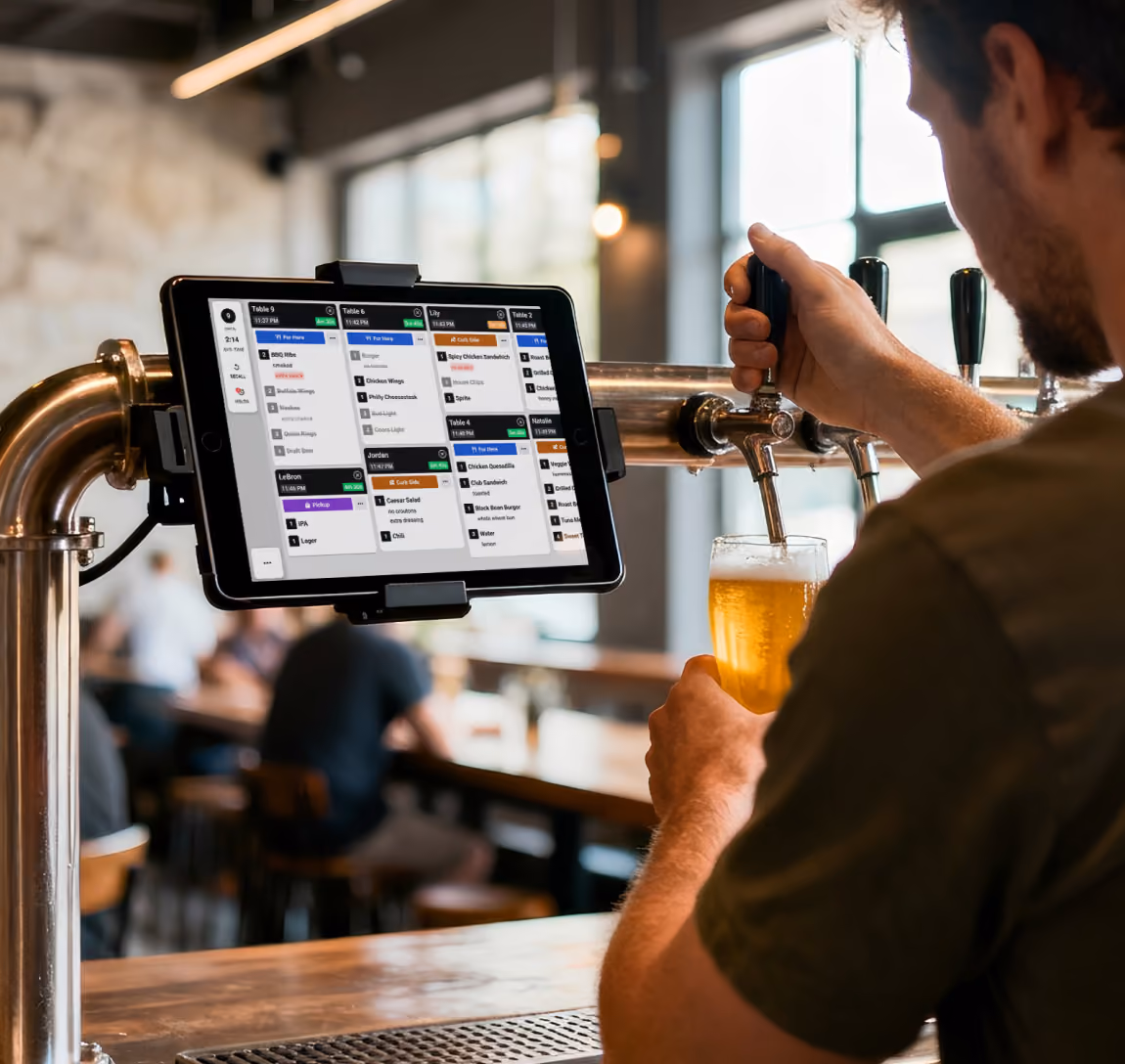 Brewery worker pouring beer from a tap, with a Fresh KDS screen displayed on a mounted tablet nearby.