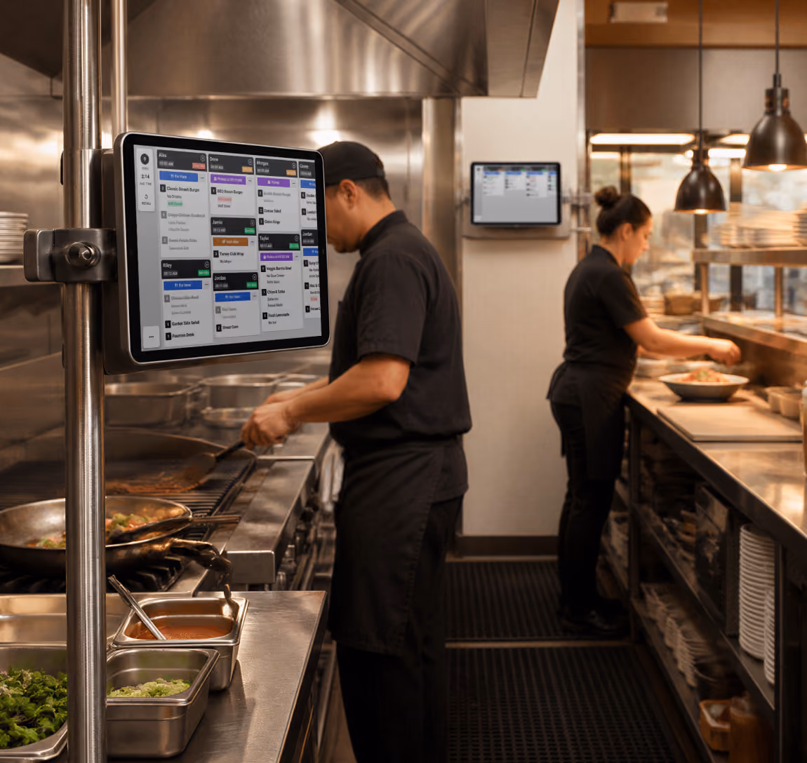Two kitchen staff preparing food in a restaurant kitchen, with Fresh KDS screens displaying orders in front of them.
