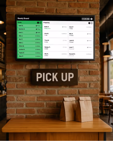 Order Tracker screen mounted above a brick wall with a 'PICK UP' sign and two bagged orders on a wooden counter.
