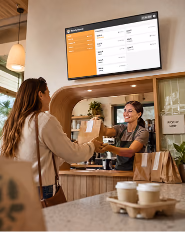 Barista handing a customer a bagged order at a café pickup counter, with drinks ready alongside.