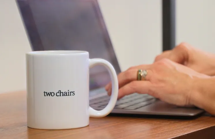 Close up of a desk with hands typing on a laptop with a nearby mug that says Two Chairs