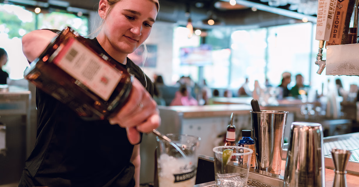 Bartender pouring liquor from a bottle into a glass with ice at a busy bar.