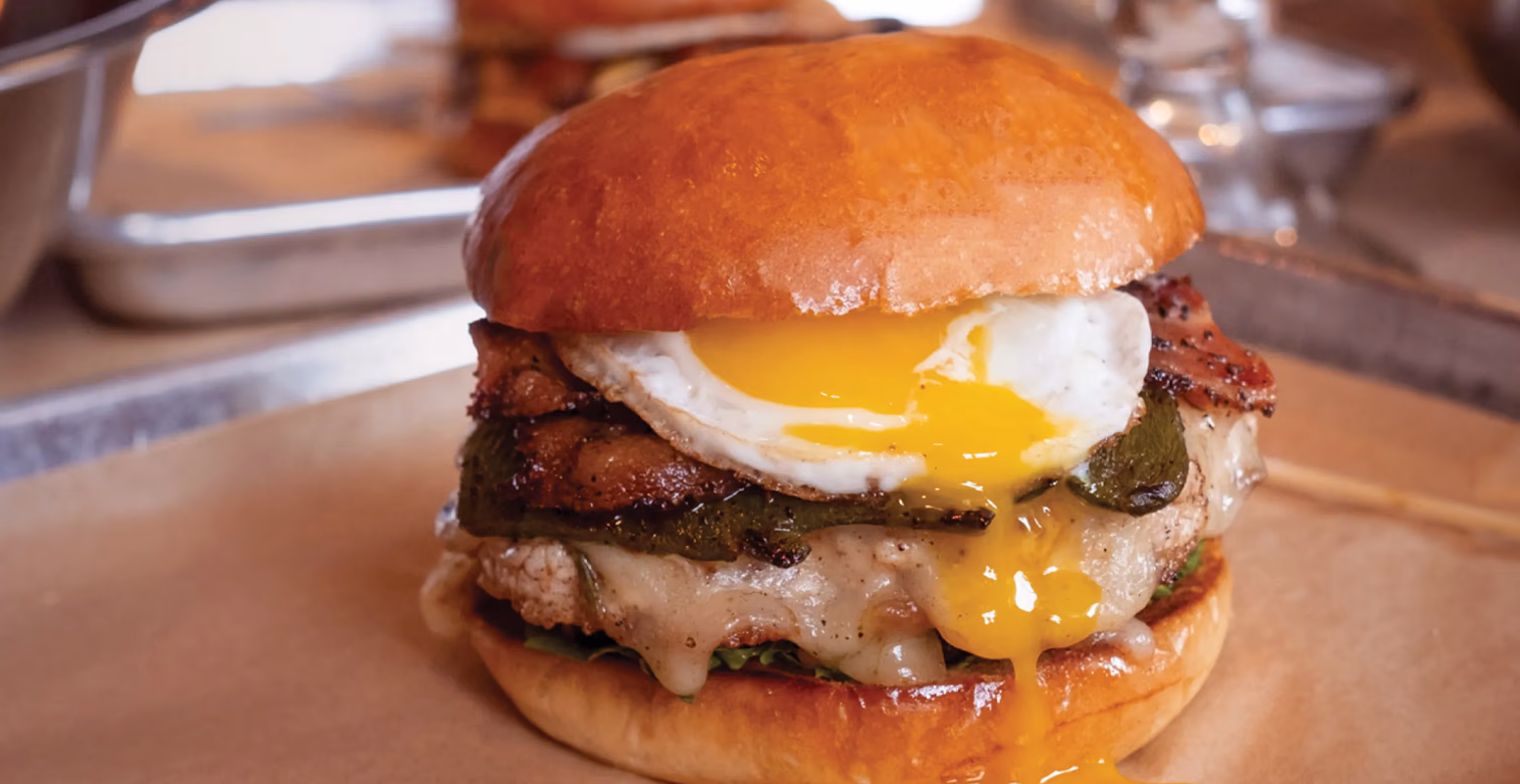 Gourmet burger with melted cheese, grilled vegetables, bacon, and a fried egg with runny yolk.