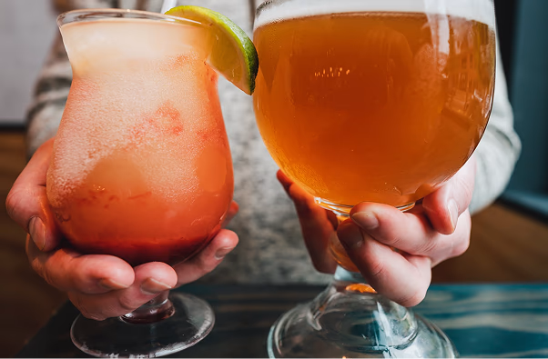 Person holding two drinks: a frosty pink cocktail with a lime wedge and a large glass of amber beer.
