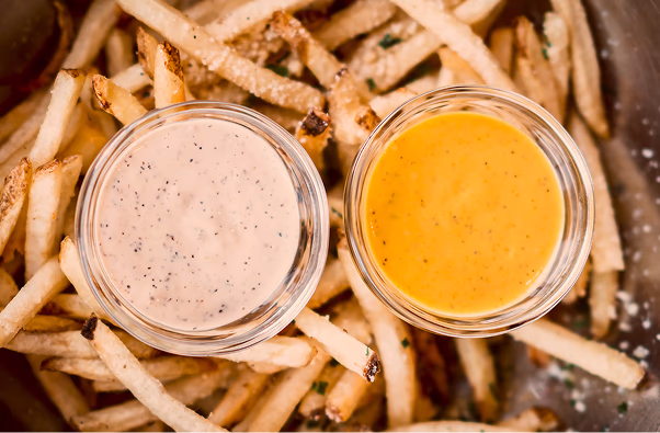 Two small glass bowls of dipping sauces, one creamy white and one orange, atop a pile of golden French fries.