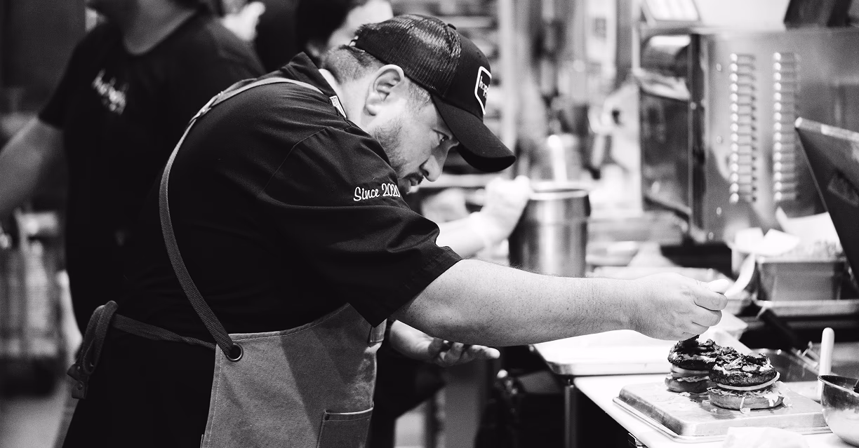 Chef in a cap and apron carefully placing toppings on stacked burgers in a busy kitchen.