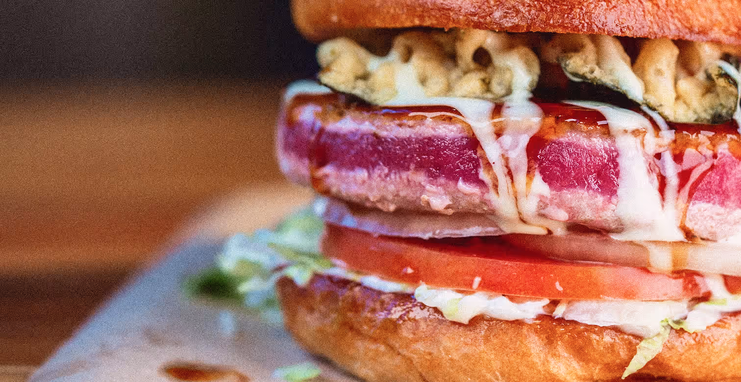 Close-up of a burger with seared tuna steak, lettuce, tomato, and sauce dripping on bun.