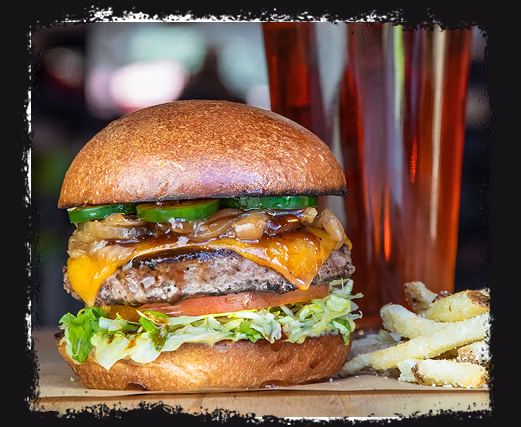 Cheeseburger with lettuce, tomato, grilled onions, jalapeños, and fries on wooden surface.