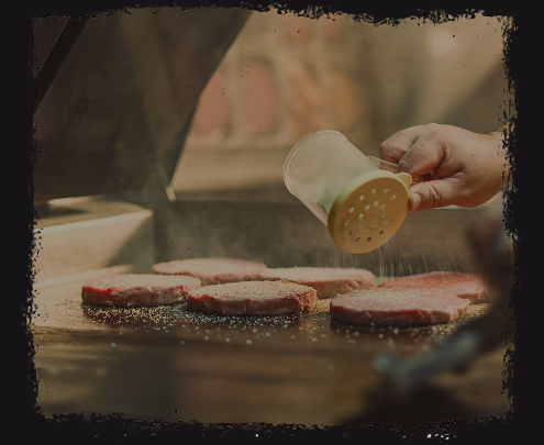 Hand sprinkling seasoning on raw beef patties cooking on a grill.