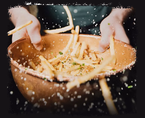 Hands tossing seasoned French fries in a bowl with salt and herbs visible in motion.