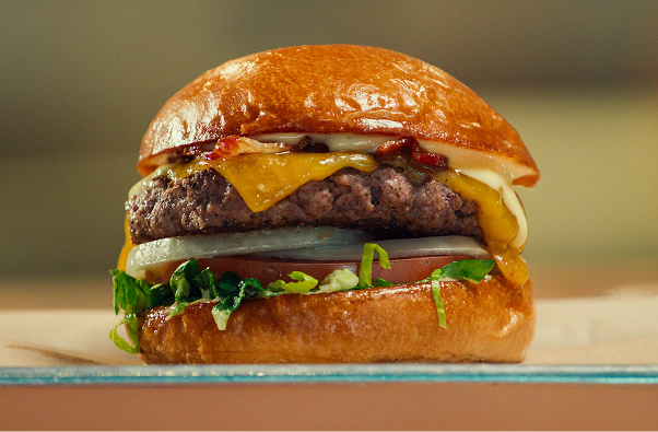 Close-up of a cheeseburger with melted cheddar cheese, crispy bacon, lettuce, tomato, onion, and creamy sauce.