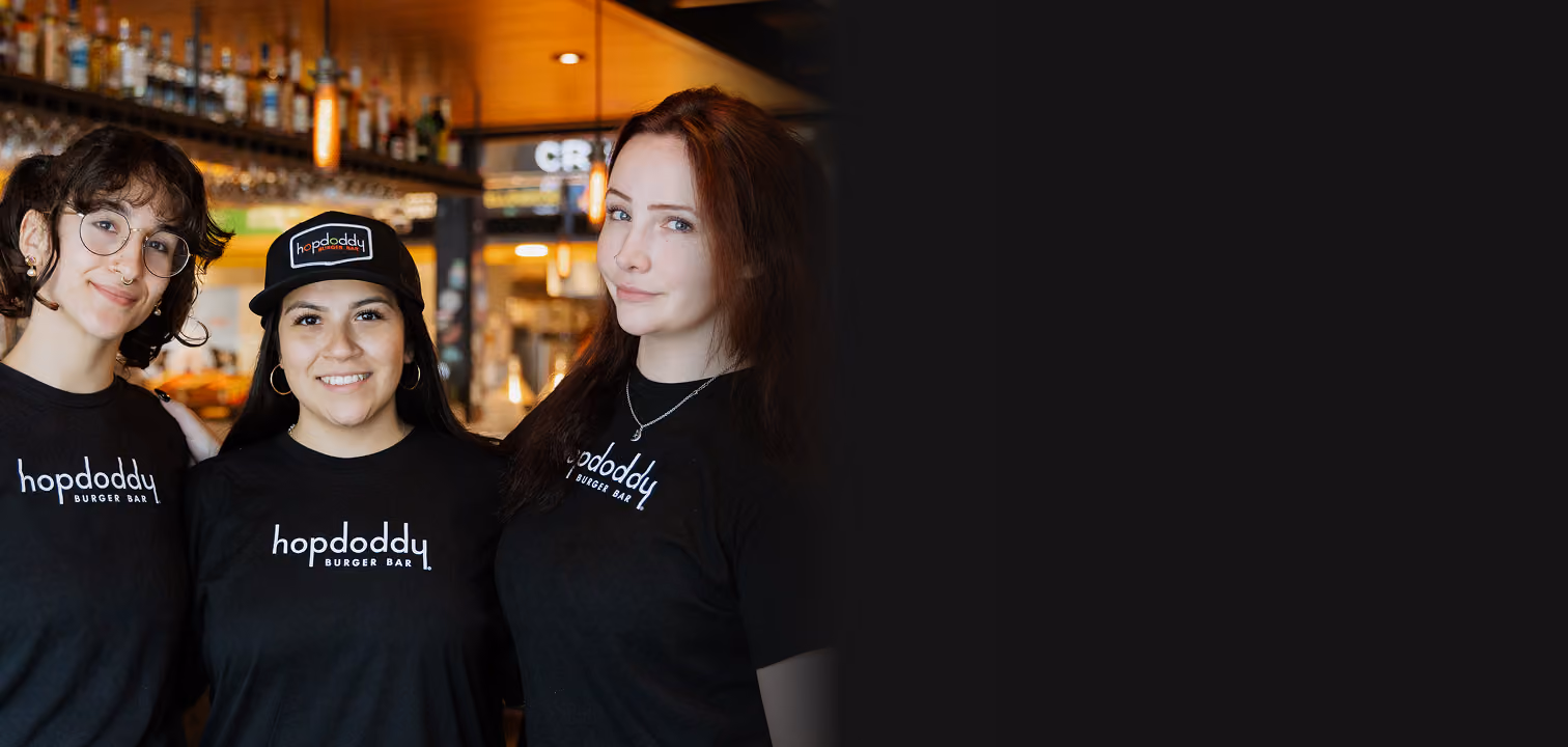 Three smiling women in Hopdoddy Burger Bar shirts posing inside a warmly lit restaurant.