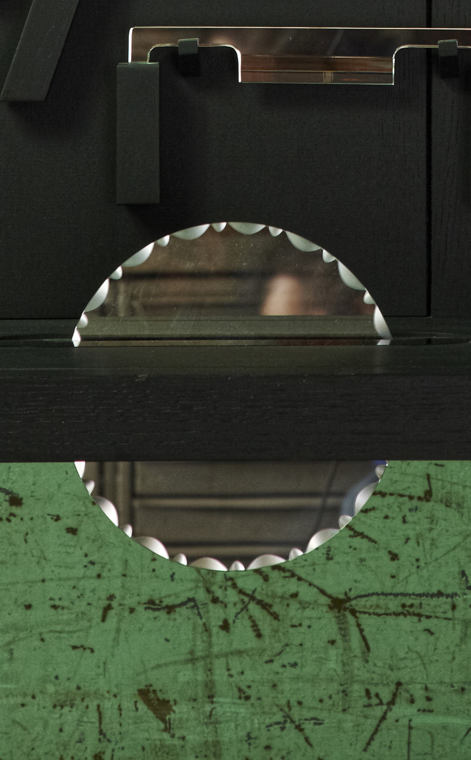 M1, détail vertical : le demi-disque de scie en miroir posé sur l'établi, sous un serre-joint en miroir ; sous le plateau, la découpe du disque laisse voir la paroi verte écaillée du monte-charge
