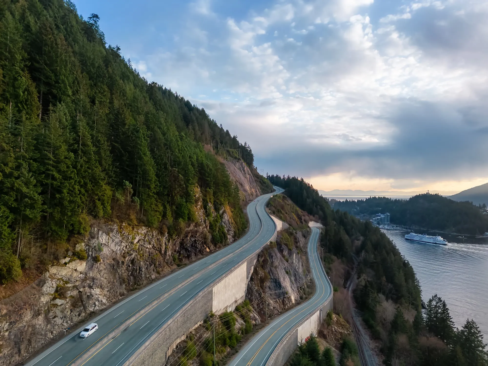 Curving highway running alongside a forested hillside with a ferry boat sailing on water in the background under a partly cloudy sky.