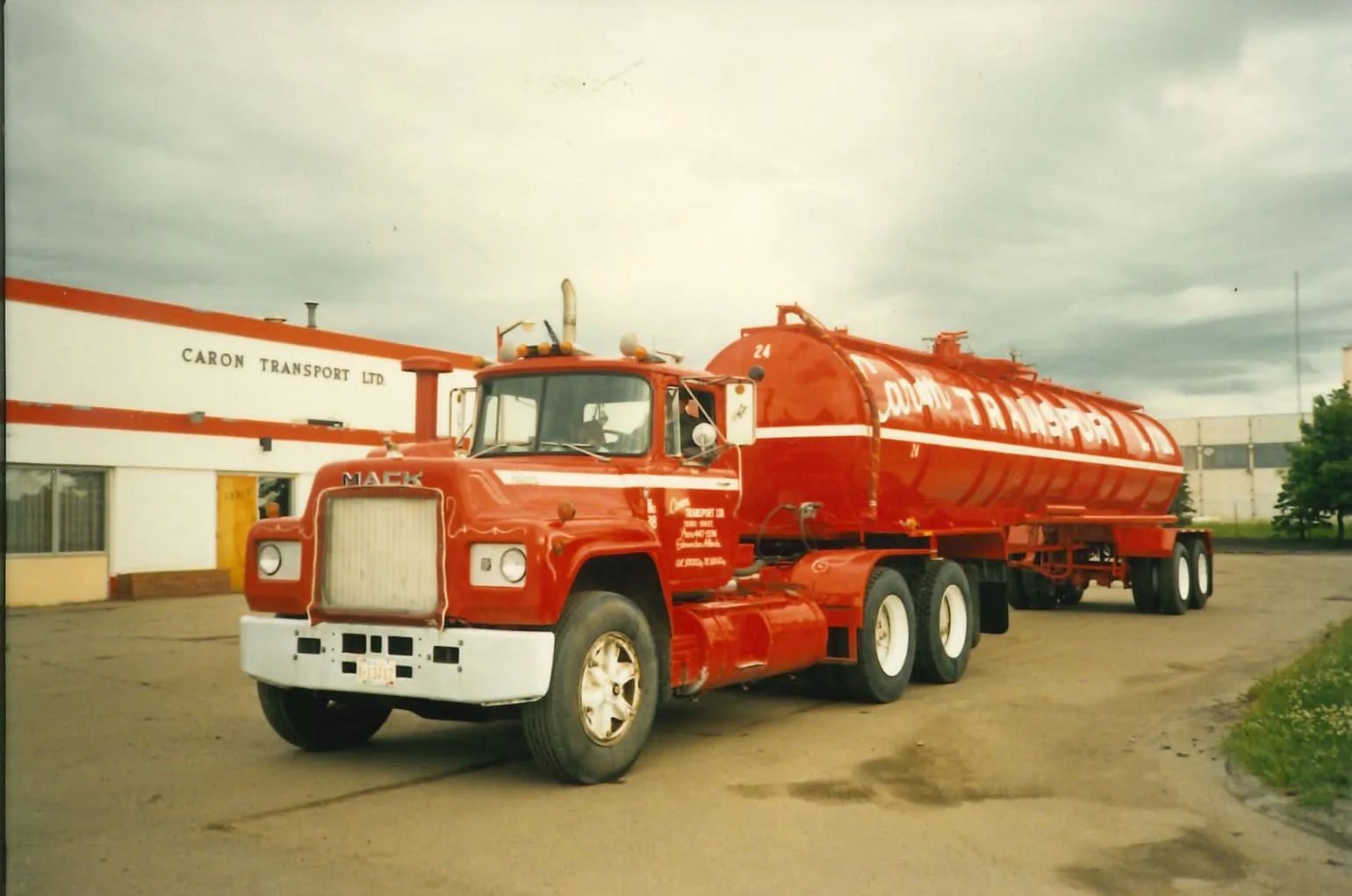 Red Mack tanker truck parked outside Caron Transport Ltd building on a cloudy day.