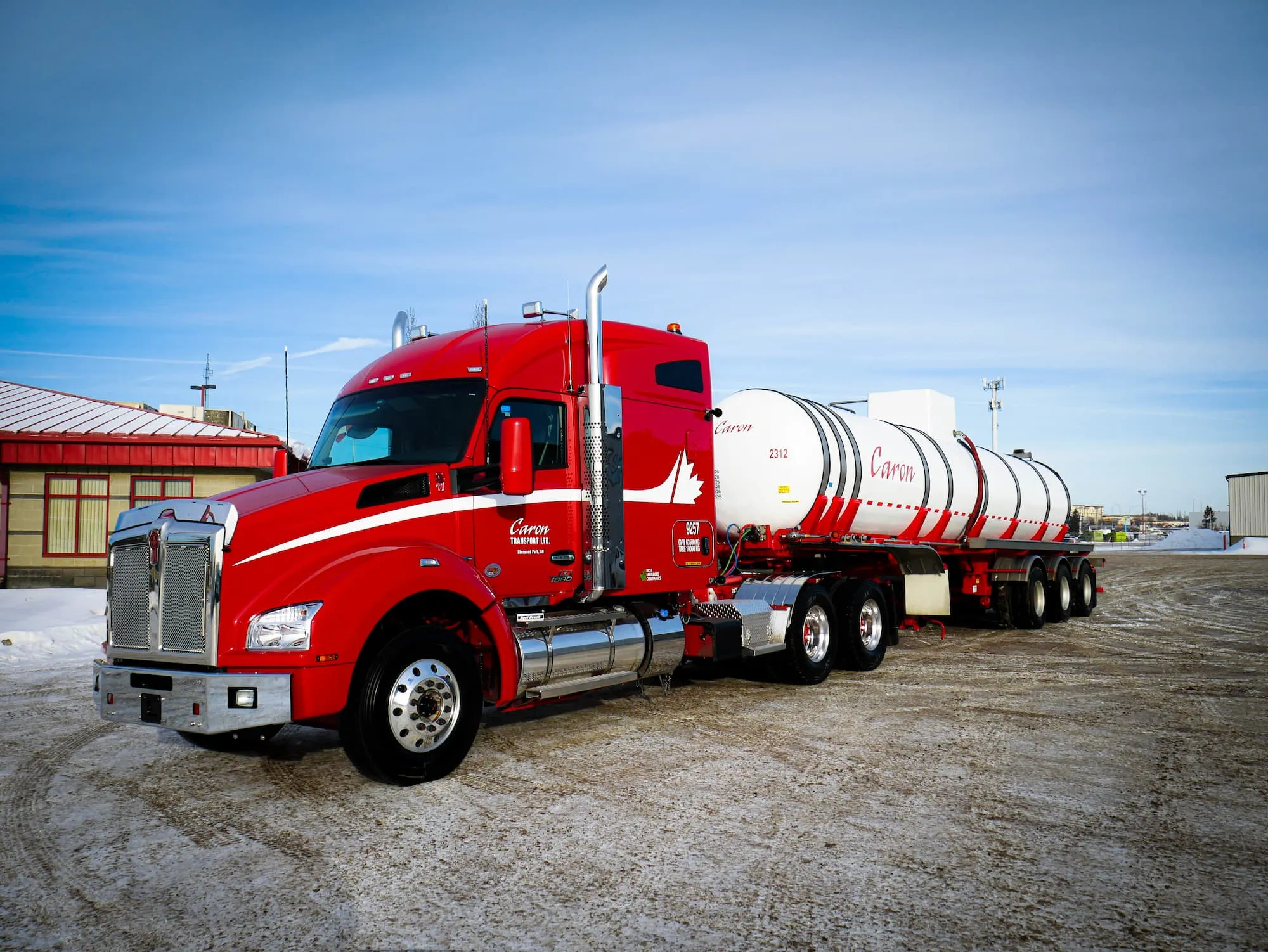 Red semi-truck with a white cylindrical tanker trailer parked on a snowy lot.