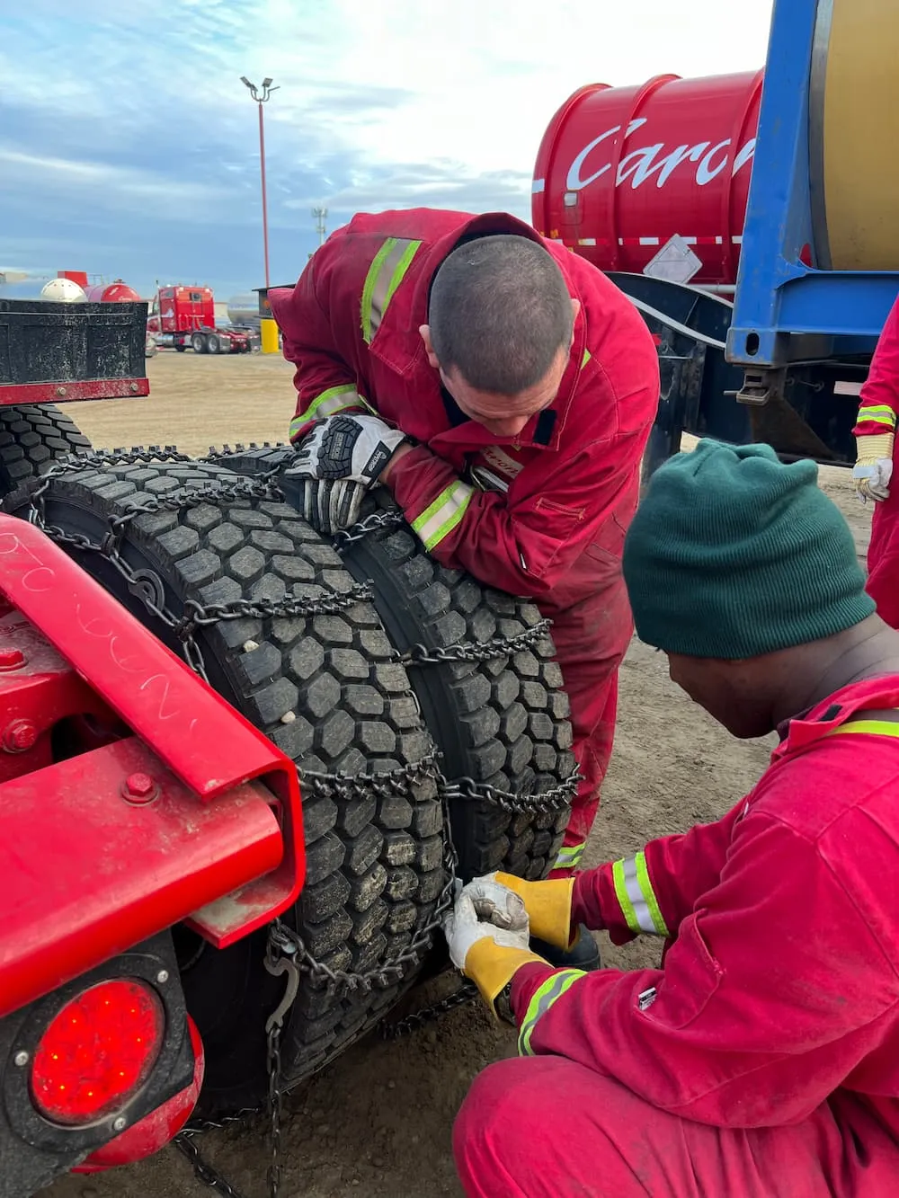 Two workers in red coveralls fitting tire chains on large truck tires.
