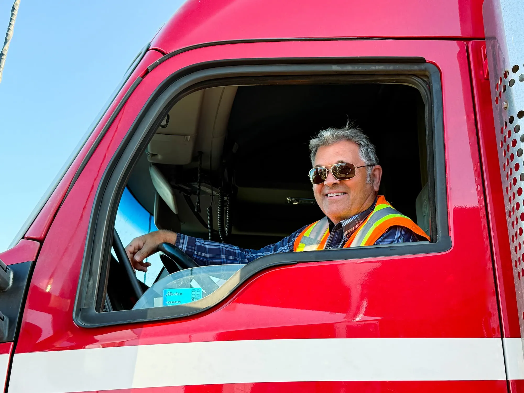 Smiling male truck driver wearing sunglasses and a safety vest sitting in the cab of a red truck.