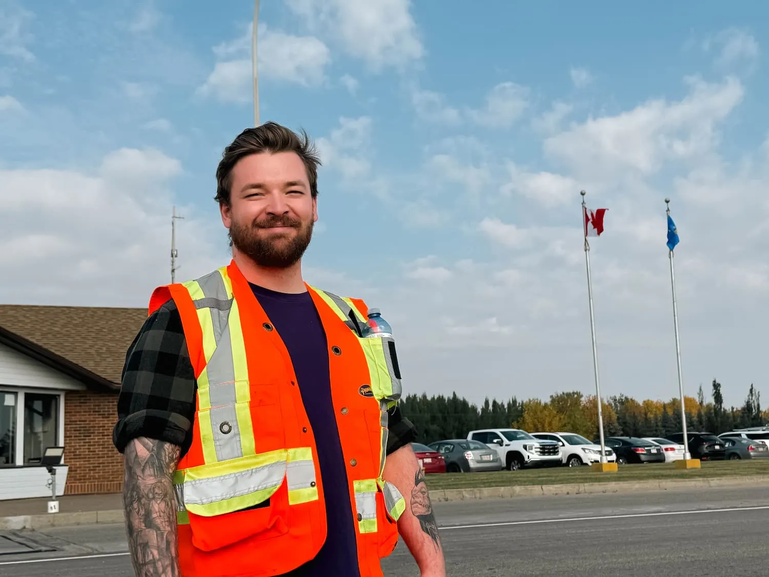 Smiling man wearing an orange reflective safety vest , standing outdoors near a building in the parking lot