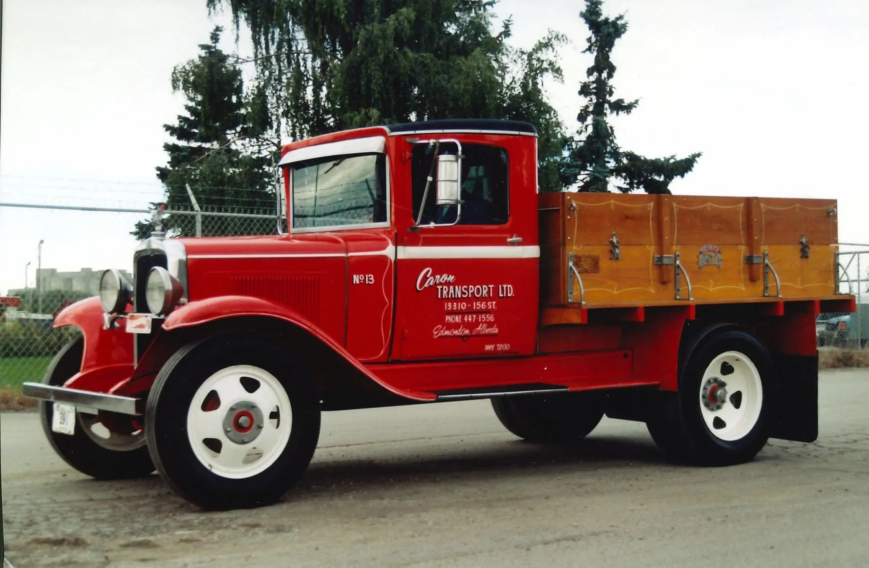 Vintage red flatbed truck with wooden side panels parked on a street with trees and fence in the background.