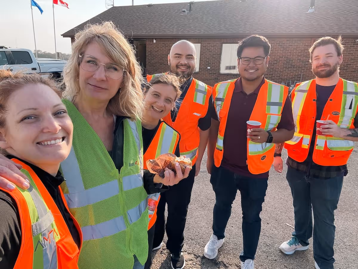 Six smiling construction workers wearing safety vests standing outside near a brick building.