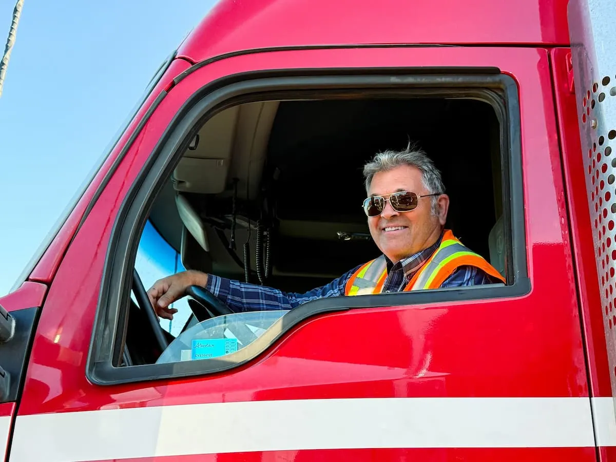 Smiling middle-aged man wearing sunglasses and a high-visibility vest sitting in the driver's seat of a red truck.