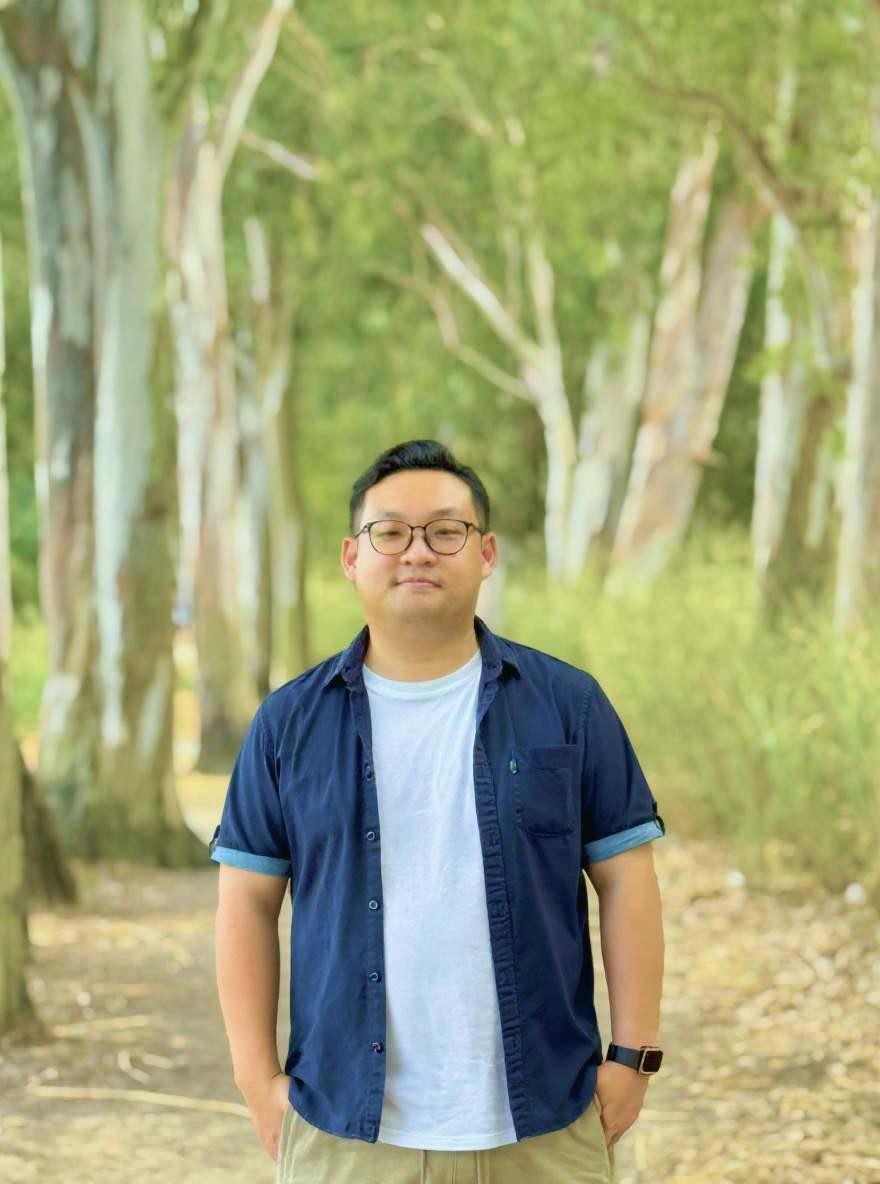 Man wearing glasses, a navy shirt over a white t-shirt, and khaki pants standing on a wooded path with trees and greenery in the background.