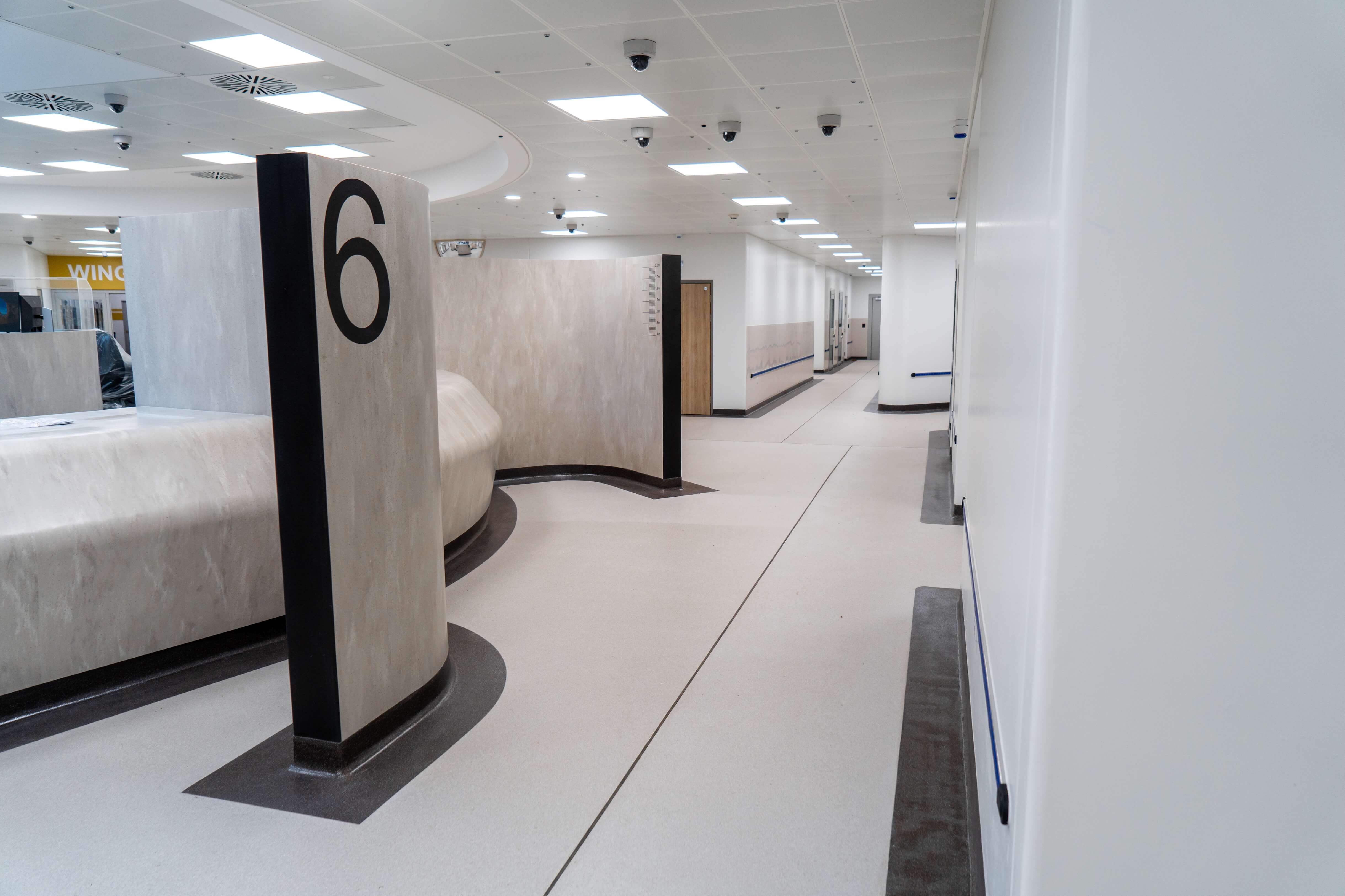 Modern, clean hallway in a building with a large numbered pillar marked 6 and surveillance cameras on the ceiling.