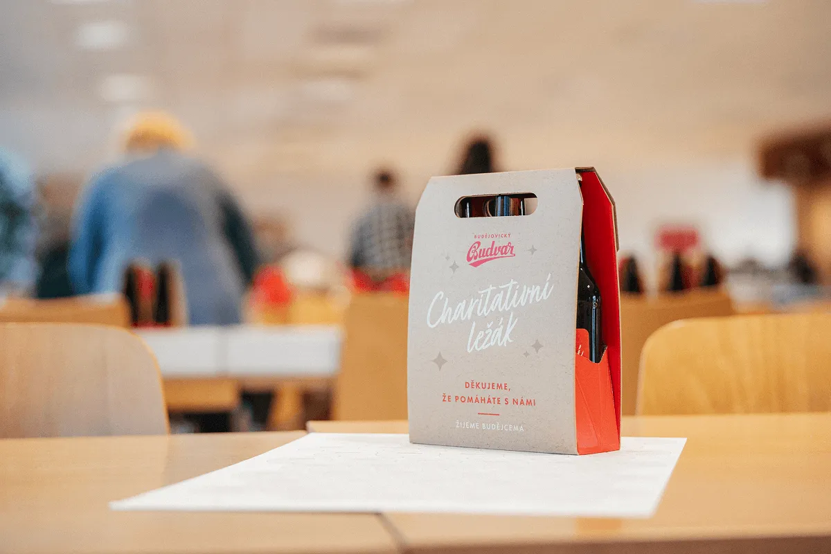 Cardboard carrier holding bottles labeled 'Budvar Charitativní ležák' on a table in a blurred indoor setting.