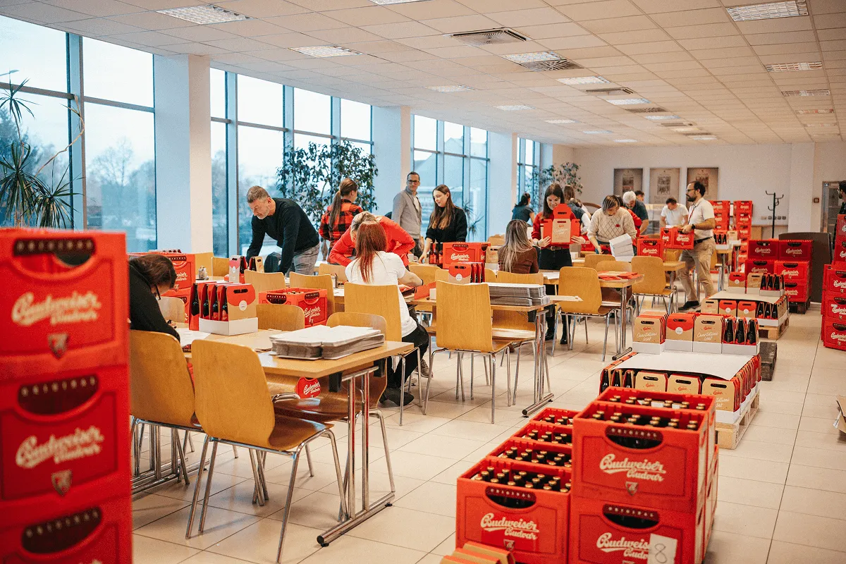 A busy room with people packing Budweiser outdoor beer bottles into red cases at wooden tables.