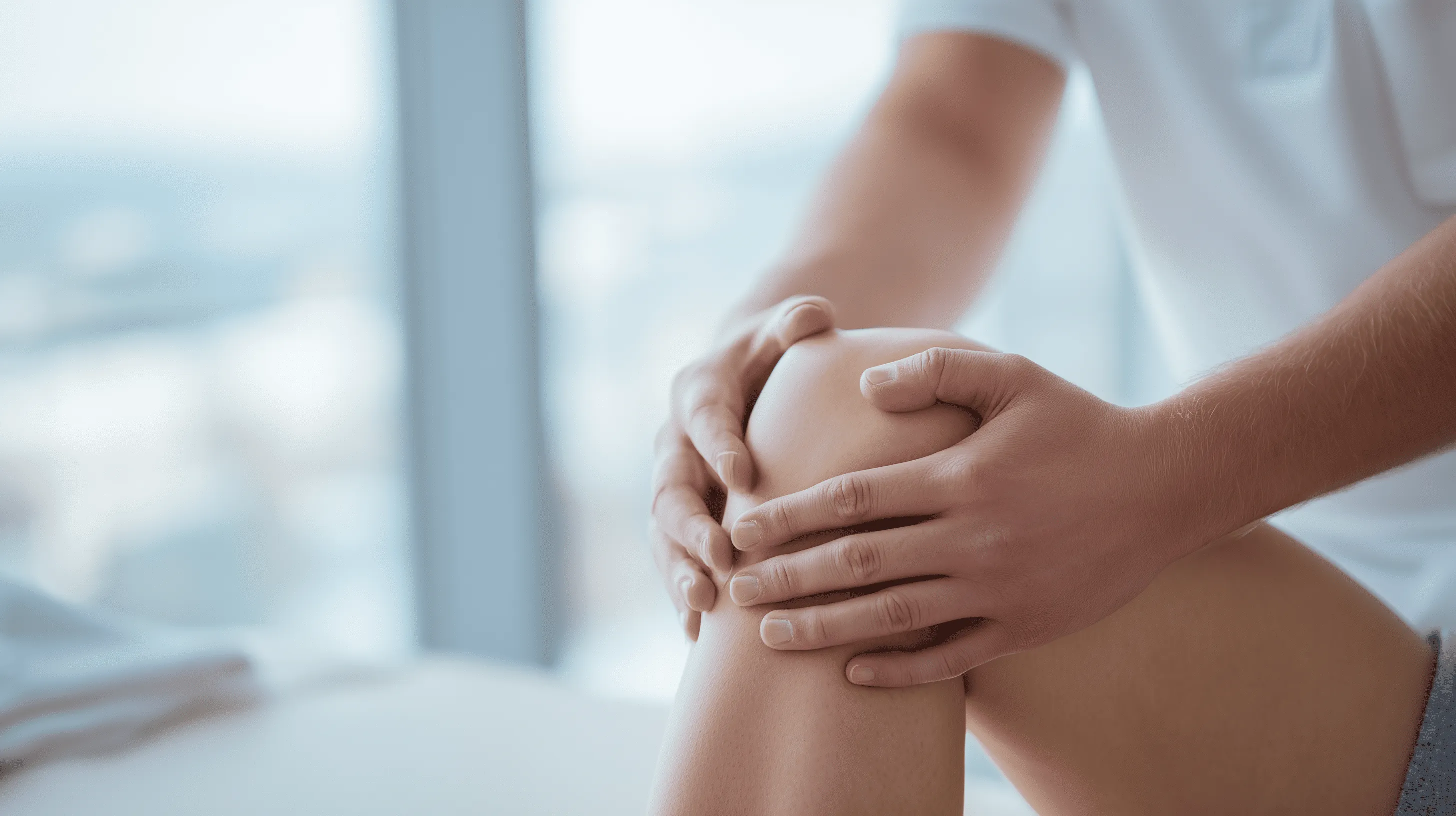 Detailaufnahme von Händen, die ein Knie halten, möglicherweise um Knieschmerzen zu lindern oder als Moment der Selbstuntersuchung nach einer Knieverletzung. Das Bild symbolisiert die Symptome und die Sorge vor einem Kreuzbandriss oder einer Kreuzbandruptur.