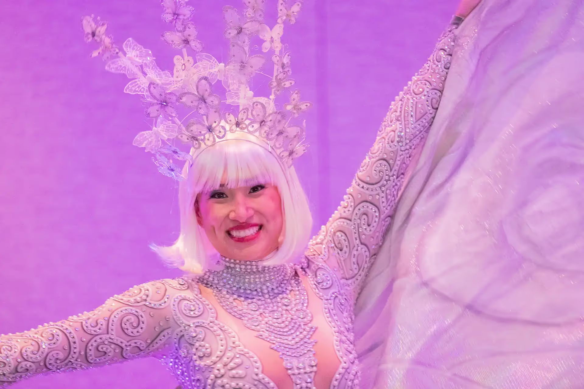 Smiling performer in a white ornate costume with pearl details and a headpiece featuring transparent butterflies against a purple background.